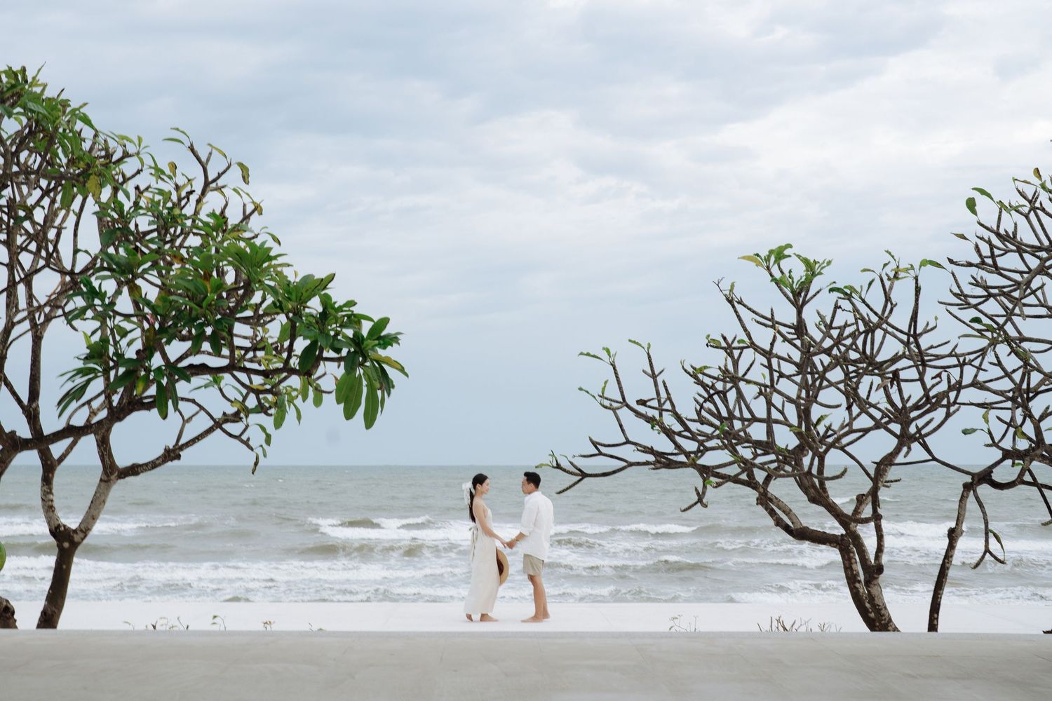 Couple in white holding hands on tropical beach between bare frangipani trees with ocean waves.