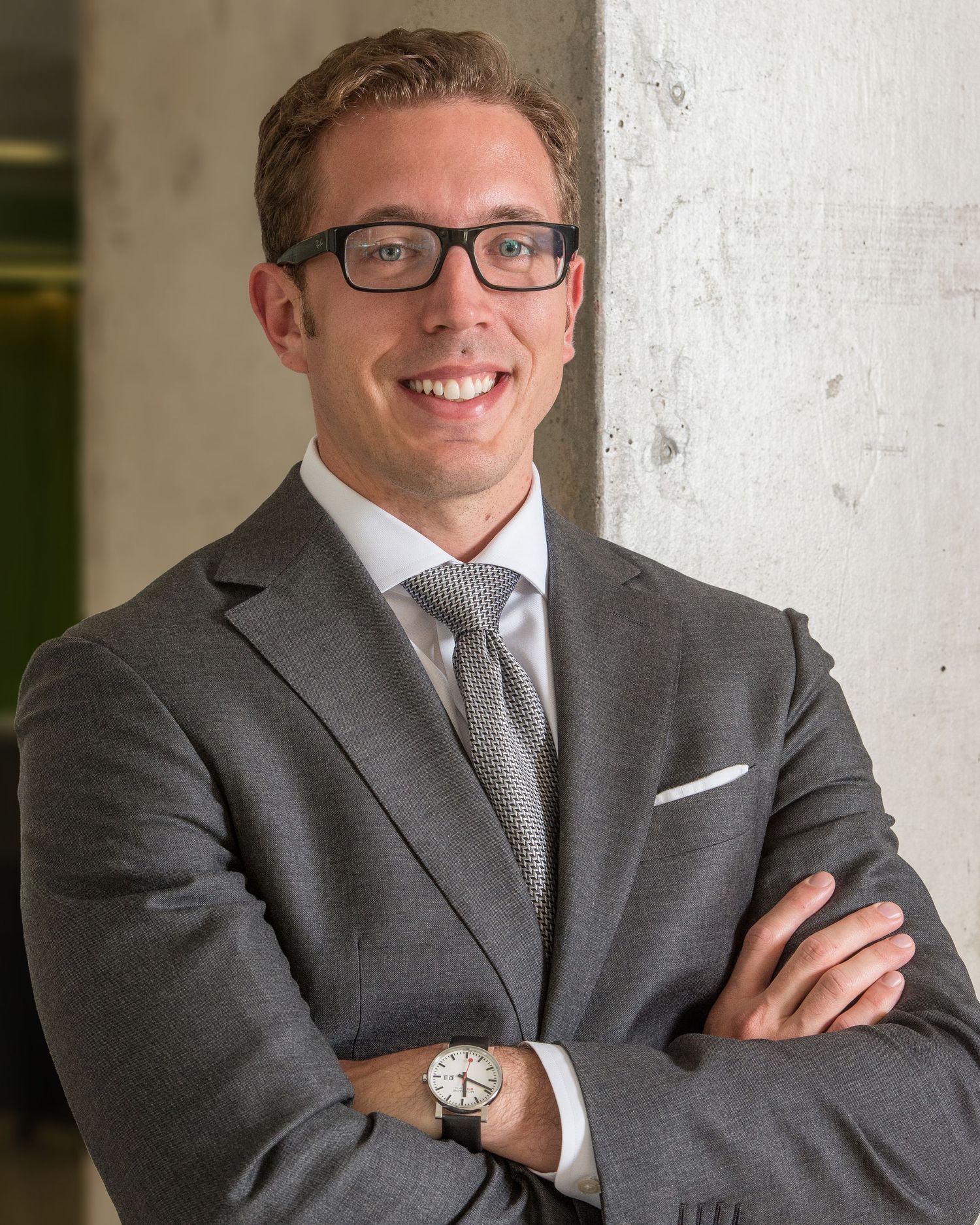 A Denver Professional posing for a professional headshot wearing a grey suit and glasses smiles confidently with arms crossed.