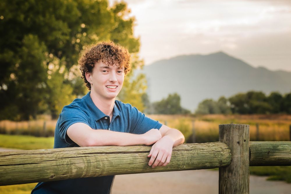 A Denver Senior in a blue shirt leans on a wooden fence with mountains and trees in the background at sunset.