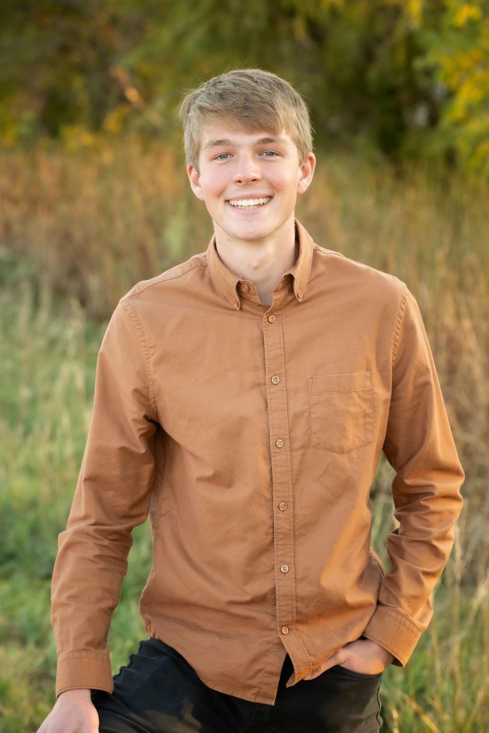 A Thornton Colorado Senior in a brown button-up shirt poses outdoors against a natural background with yellow wildflowers.