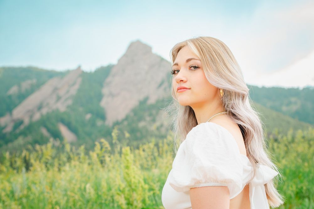 A Thornton Colorado Senior in a white dress stands in a mountain meadow with sunlit peaks and wildflowers in the background.