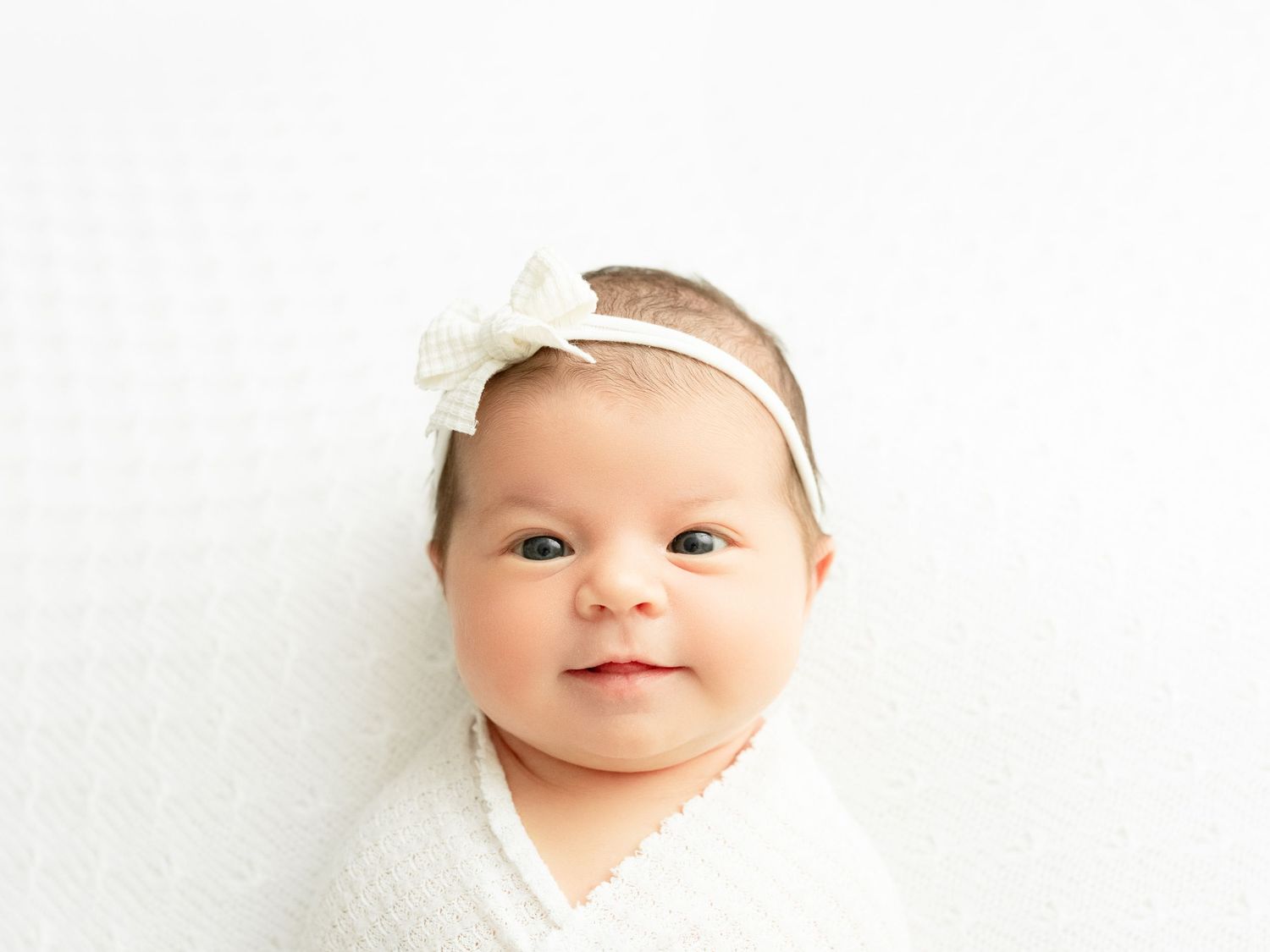 A yawning newborn baby swaddled in white fabric against a white background with a small headband.