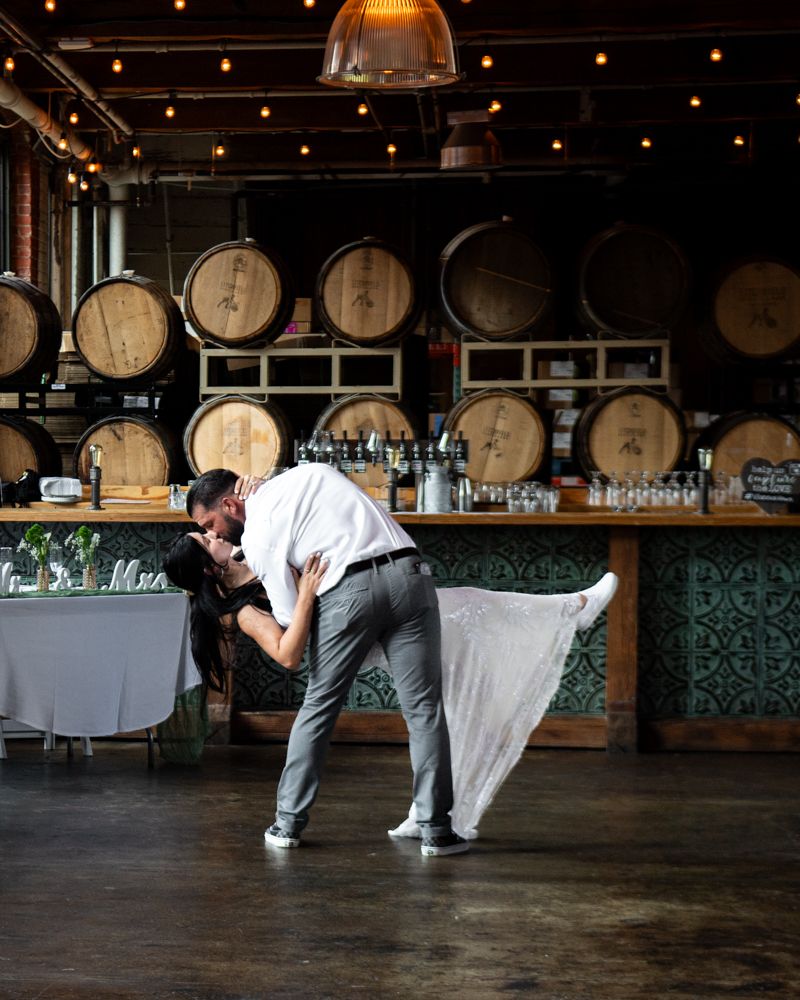 A couple performs a choreographed dance move in a rustic venue with wooden barrels in the background.