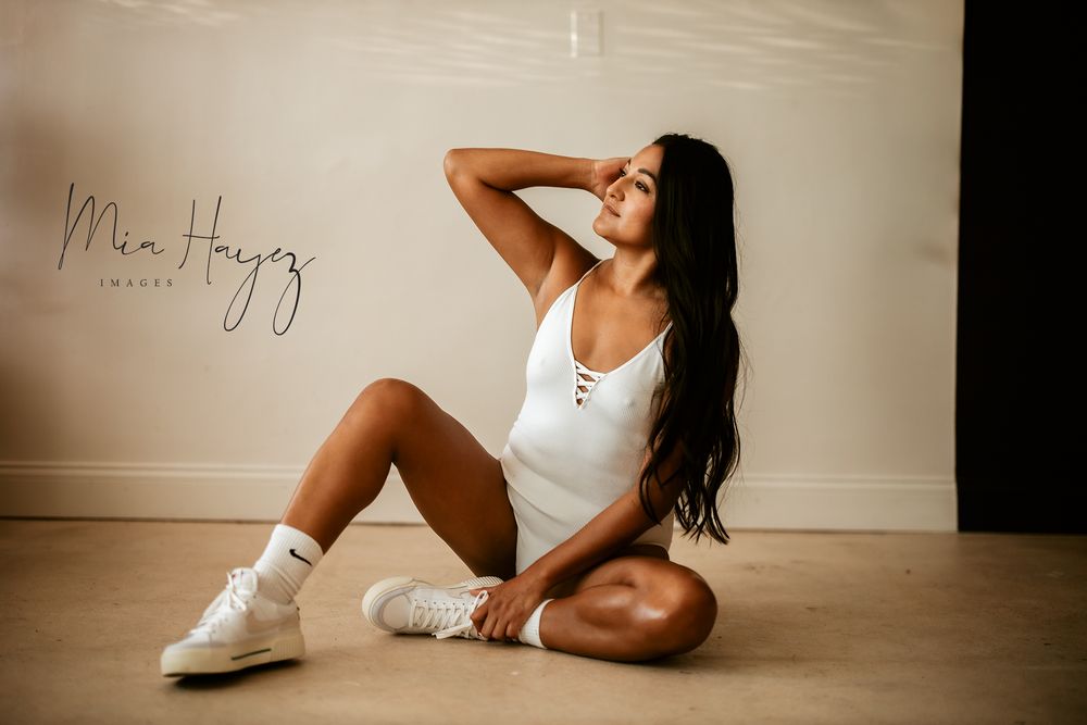 Athletic portrait in white bodysuit and sneakers against neutral wall.