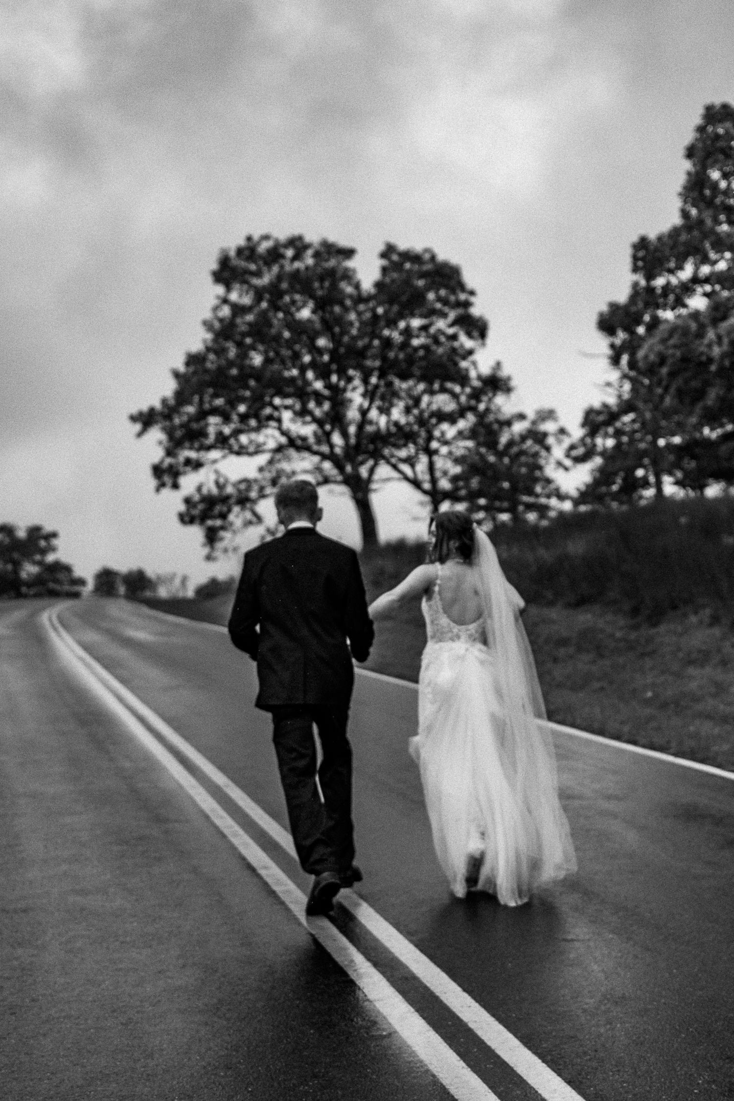 bride and groom running away from camera in the road with trees behind them.
