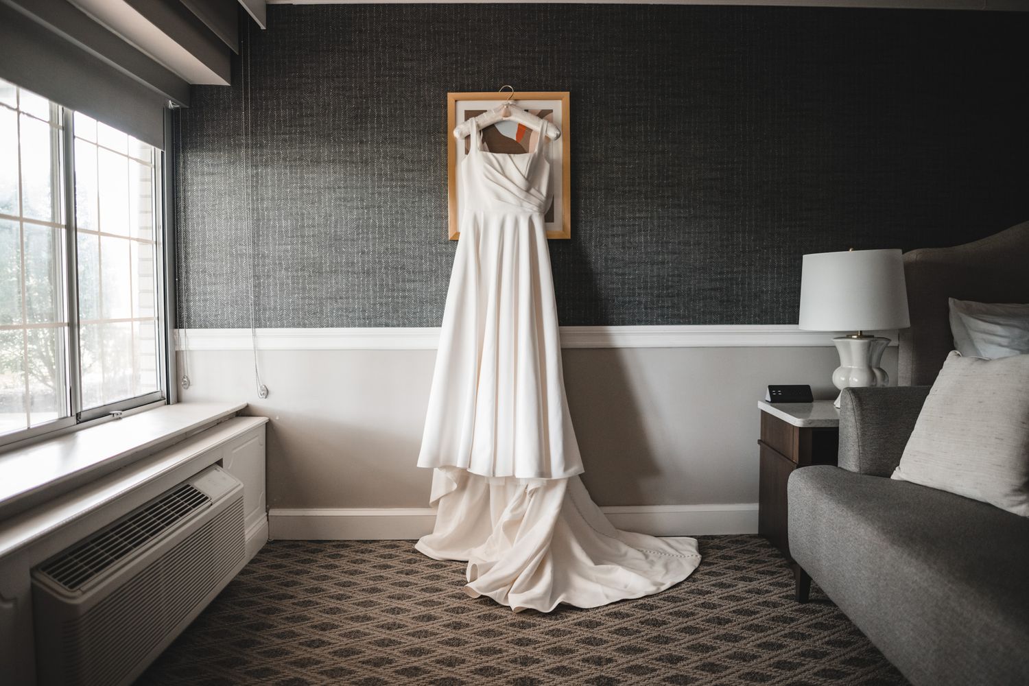 An elegant white wedding dress hangs on a wooden hanger against a gray wall with natural window light.