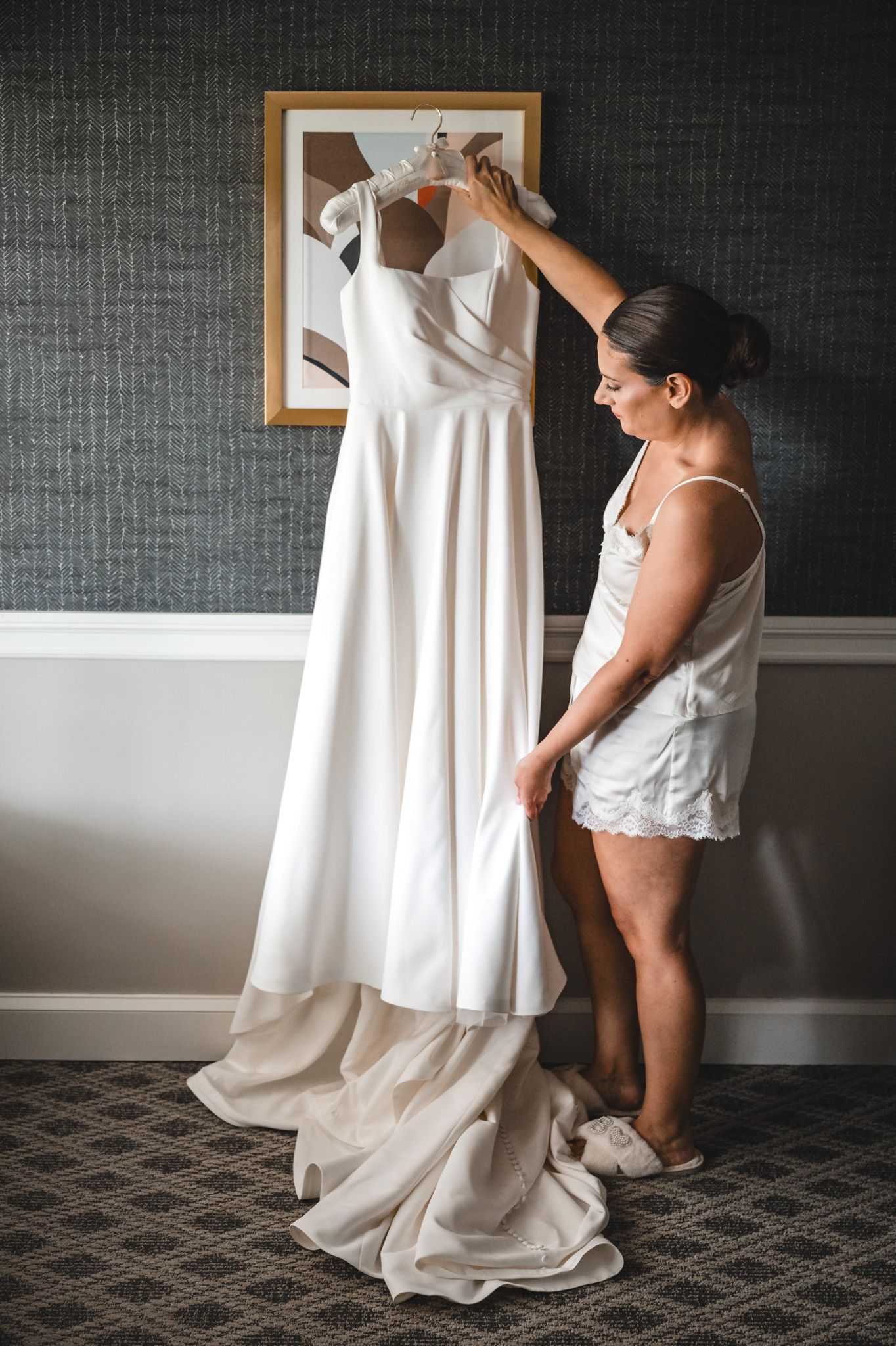 A white wedding dress being adjusted on a hanger against a textured gray wall.