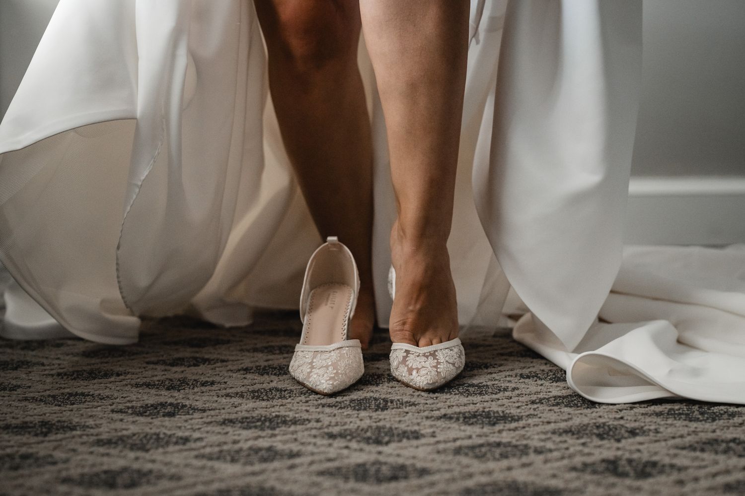 Close-up of delicate lace wedding shoes with white dress hem on gray carpet.