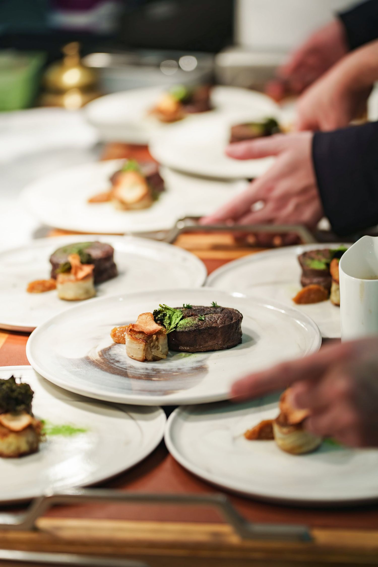 Chefs plating elegant gourmet dishes with seared meat and scallops on white plates in a professional kitchen.