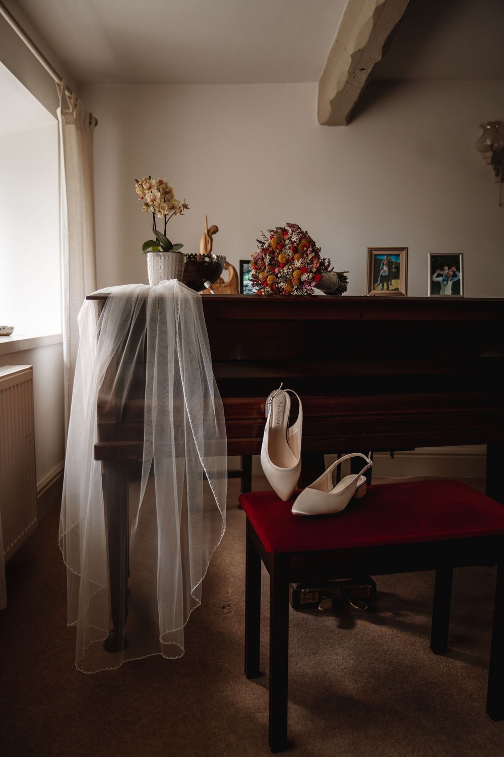 Bridal veil draped over chair beside white heels on red stool near piano.
