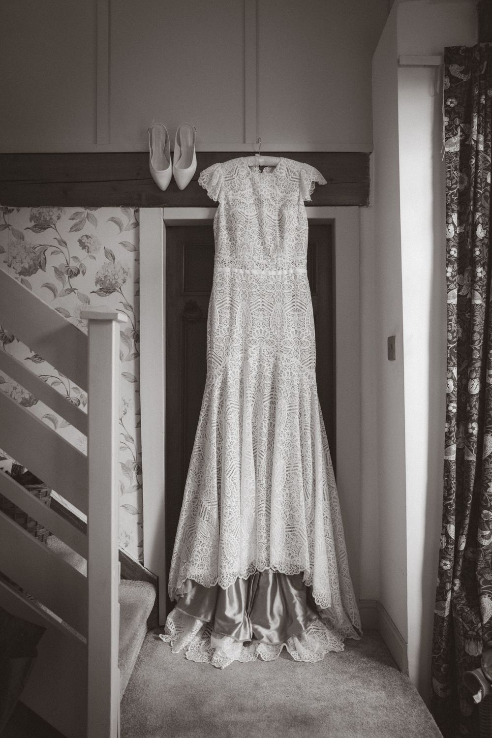 Elegant lace wedding dress hanging on a door with bridal heels displayed above in a black and white photo.