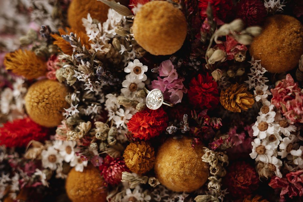 Engagement ring nestled in vibrant dried flower bouquet with red, orange and white blooms.