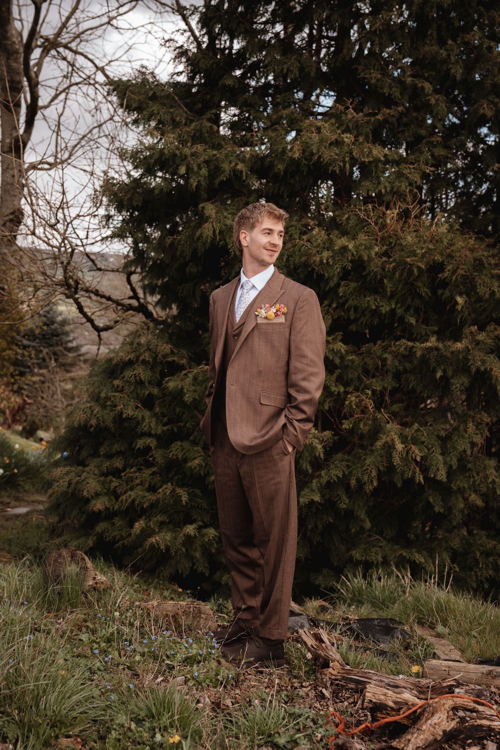 Groom in brown three-piece suit standing outdoors among evergreen trees on wedding day.