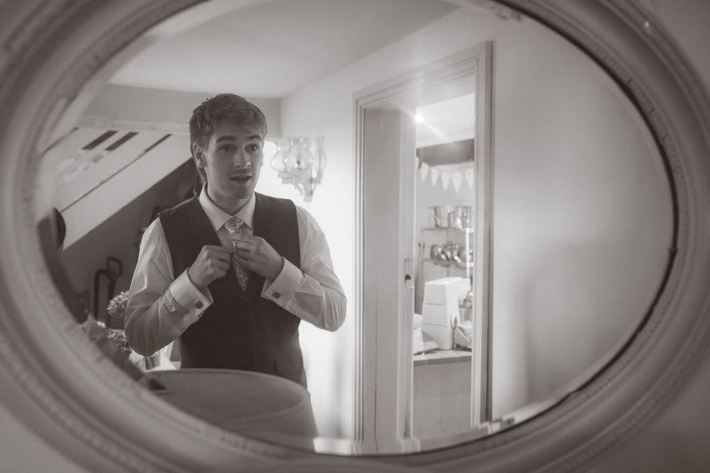 Groom adjusting his tie reflected in an oval mirror, black and white wedding preparation photo.