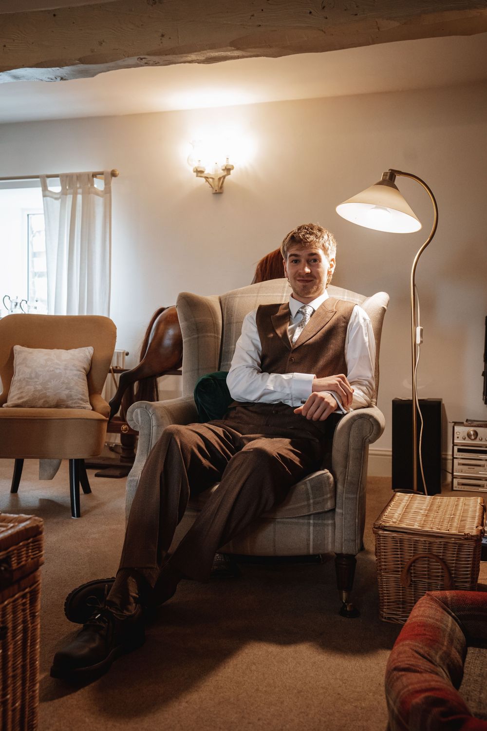 Groom in brown vest and tie sits relaxed in armchair in warmly lit rustic room before wedding.