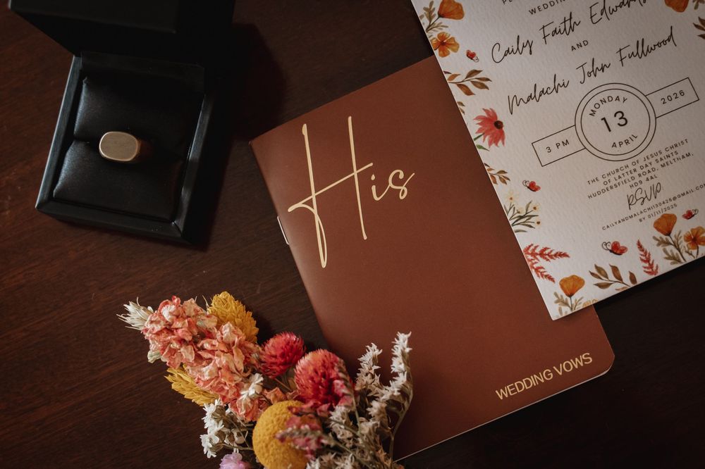 Brown leather wedding vows book labeled His beside floral invitation, dried flowers, and ring box on dark wood table.
