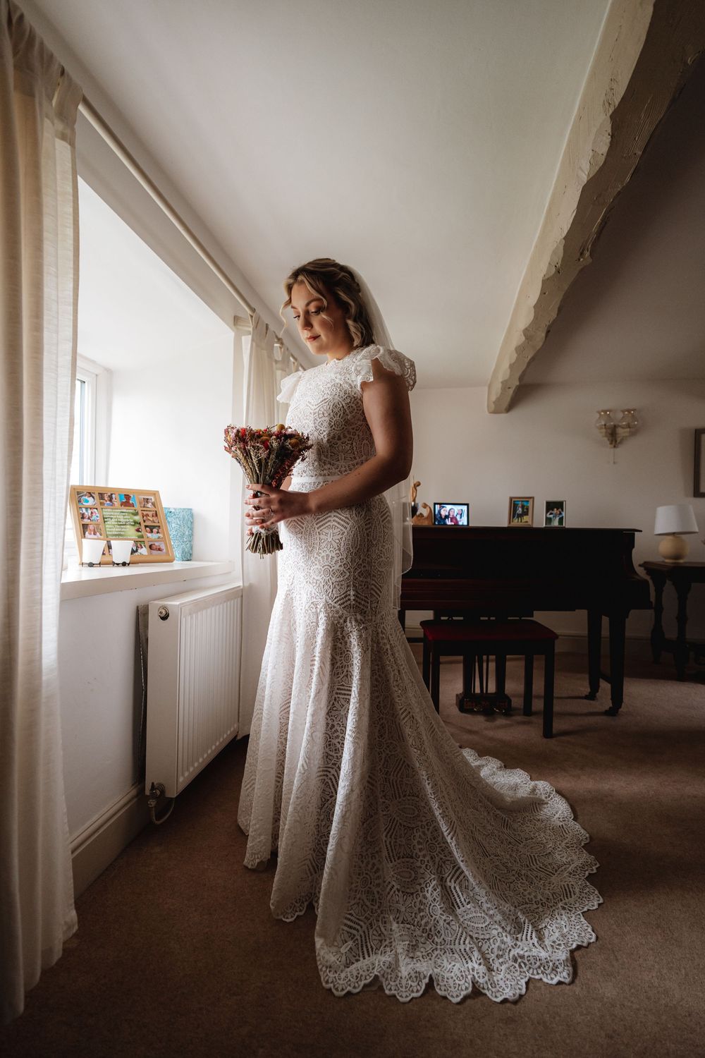 Bride in a fitted lace wedding gown holding a dried flower bouquet standing by a window in a cozy room.