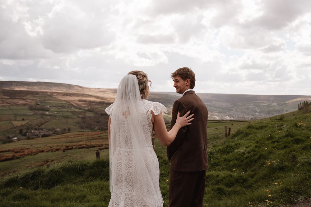 A bride and groom stand together on a lush green hillside overlooking a scenic valley on their wedding day.