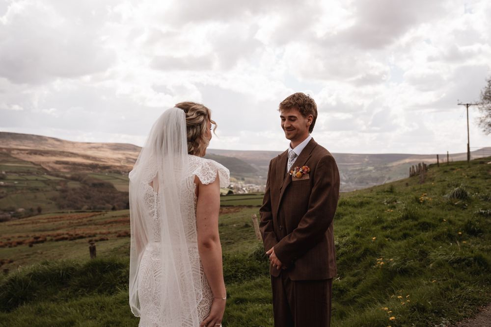 Groom smiles at bride during outdoor wedding ceremony with scenic countryside backdrop.