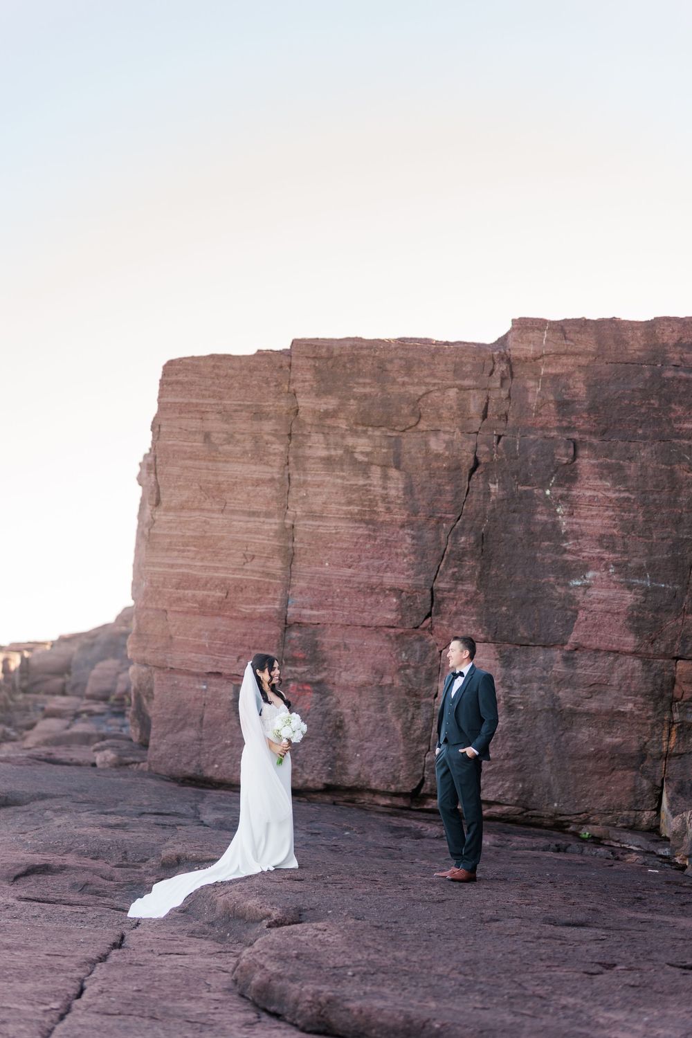 A couple poses for wedding photos against dramatic red rock cliffs at sunset in a desert landscape.