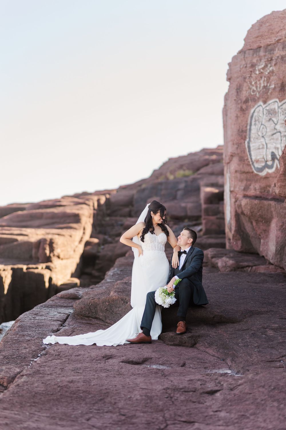 A couple in wedding attire sits together on red rock formations during a desert photo session at sunset.