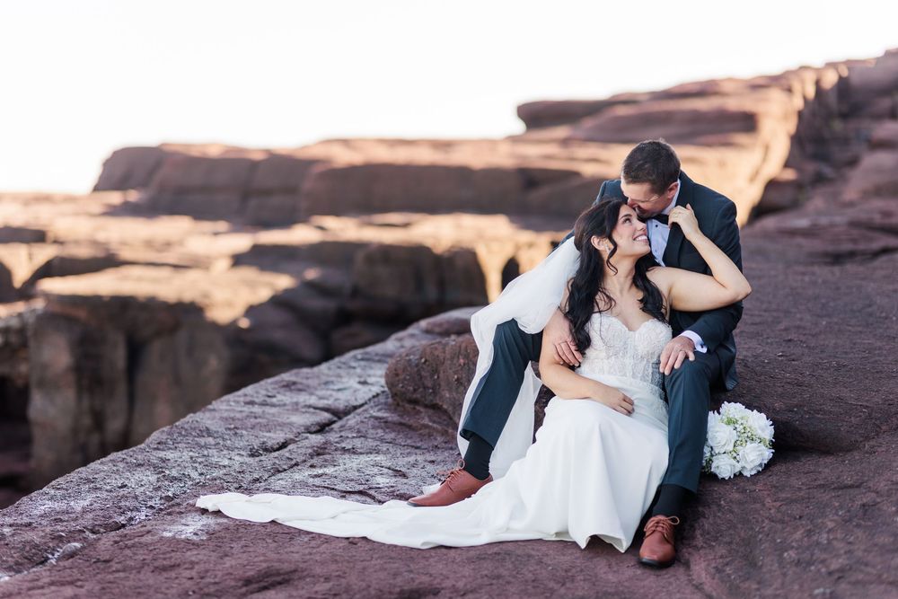 Couple in wedding attire sits together on red rock formations during sunset in desert landscape.