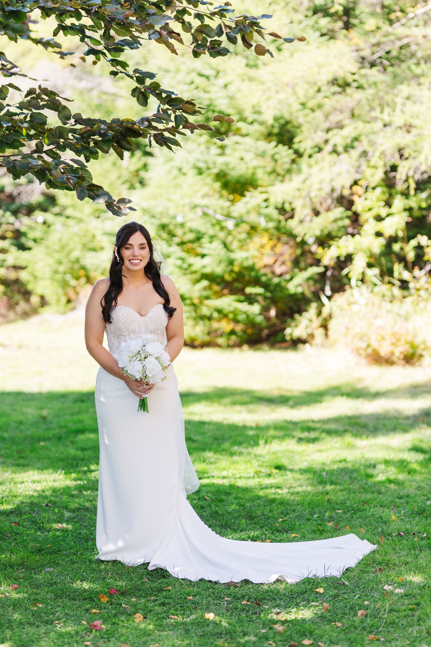 Bride in a flowing white lace wedding dress poses gracefully on a sunlit lawn surrounded by green foliage.