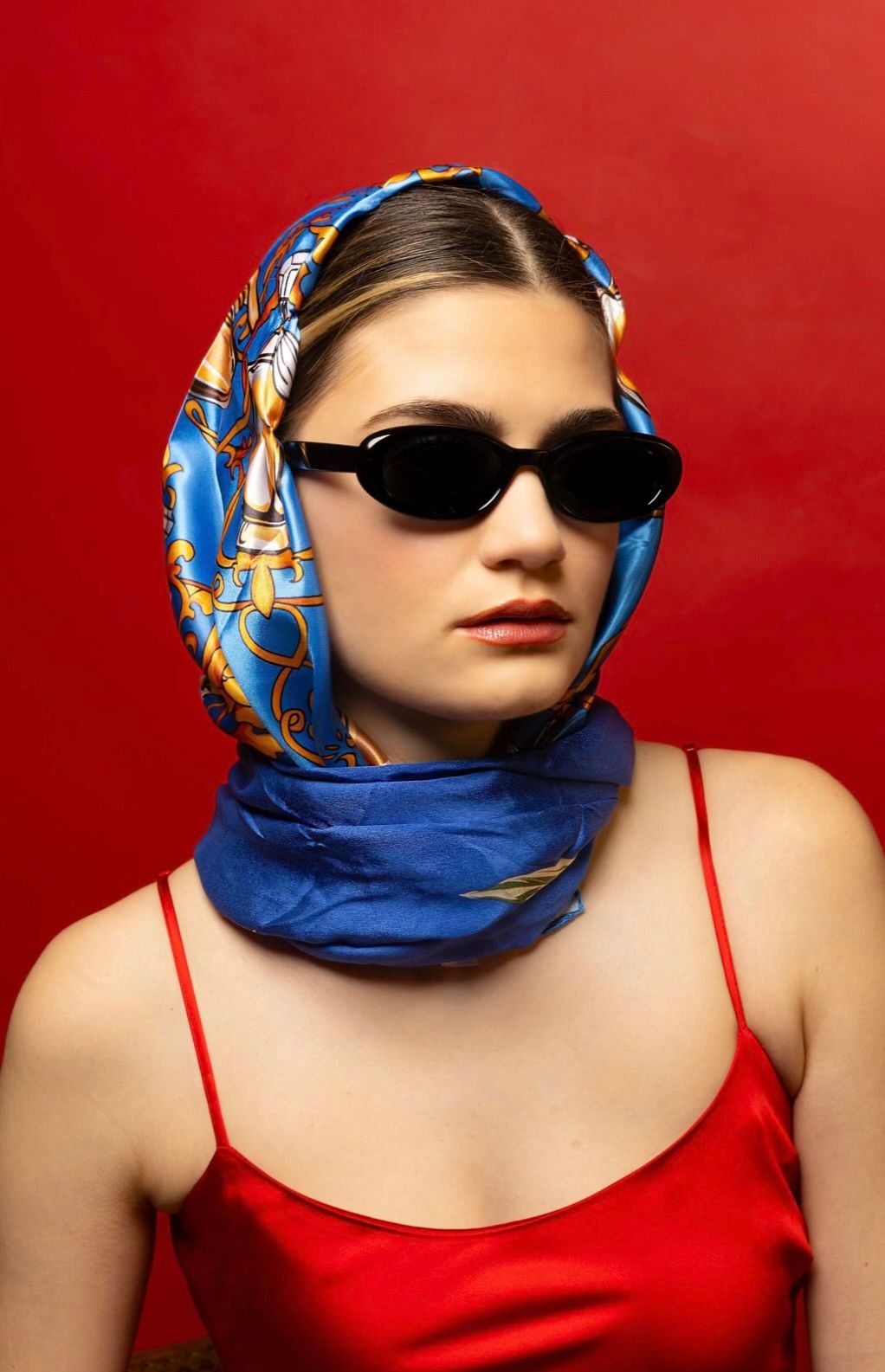 Red backdrop with a girl in a red dress and a blue head scarf, looking to the right with sunglasses on
