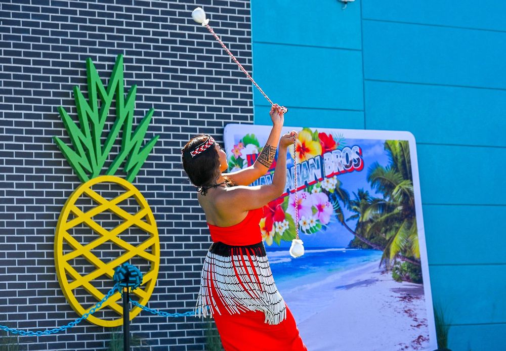 Polynesian performer spins fire poi beside large pineapple mural at Hawaiian Bros opening.