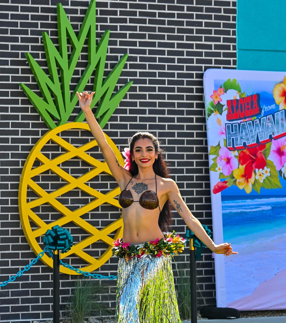Smiling hula dancer in grass skirt poses by giant pineapple prop at Aloha Hawaii themed celebration.