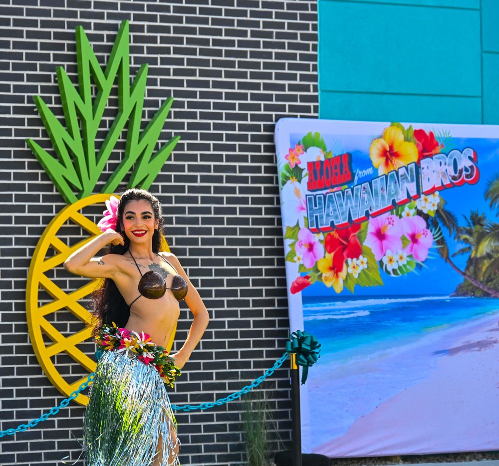 Hula dancer in grass skirt smiles beside Hawaiian Bros banner and large pineapple wall decoration.