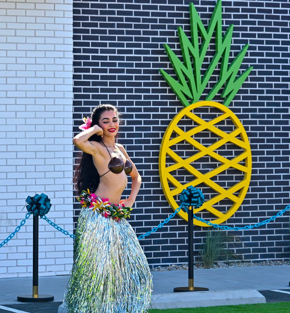 Hula dancer in grass skirt and floral accessories poses gracefully beside large pineapple art installation.