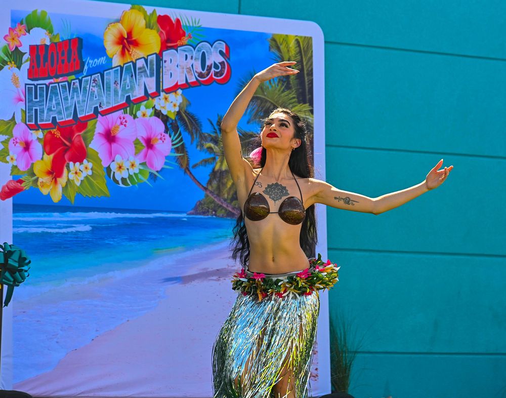 A hula dancer in a grass skirt and floral costume performs in front of a Hawaiian Bros restaurant sign.