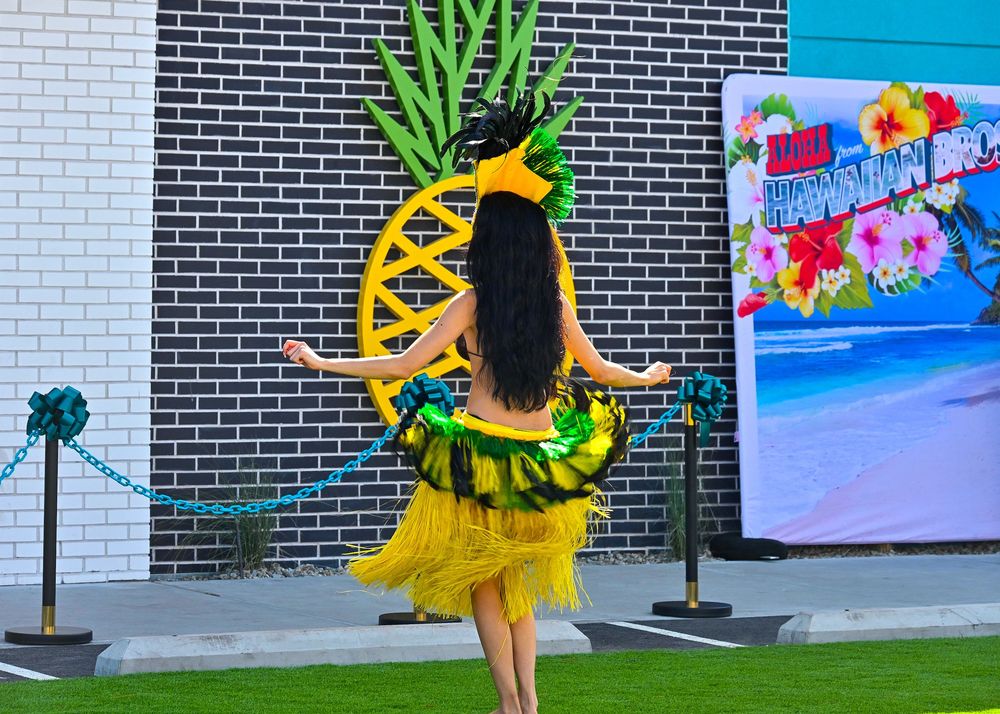 Hula dancer in yellow grass skirt performs in front of large pineapple decoration at Aloha Hawaiian Bros event.