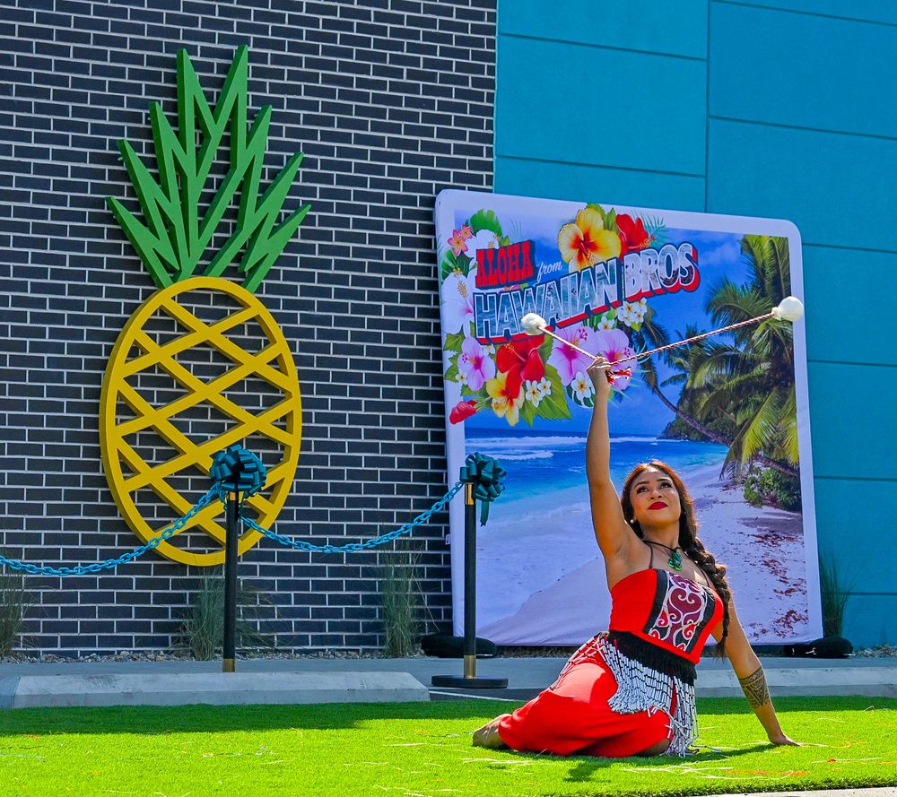Hula dancer in red outfit sits gracefully on grass beside large pineapple decoration at Hawaiian event.