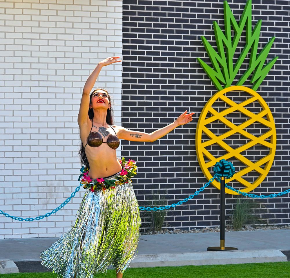Hula dancer in grass skirt and floral lei performs graceful pose beside large pineapple decoration.