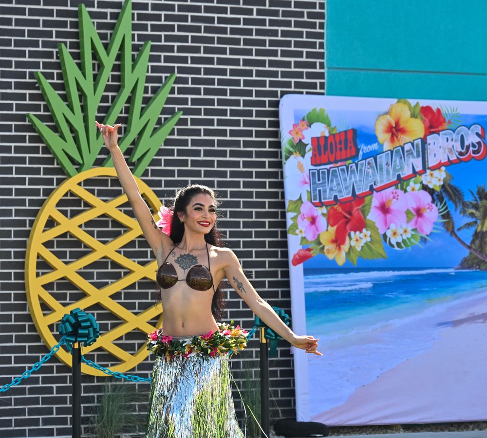 Hula dancer in green grass skirt performs beside decorative pineapple at Hawaiian Bros Island Grill event.