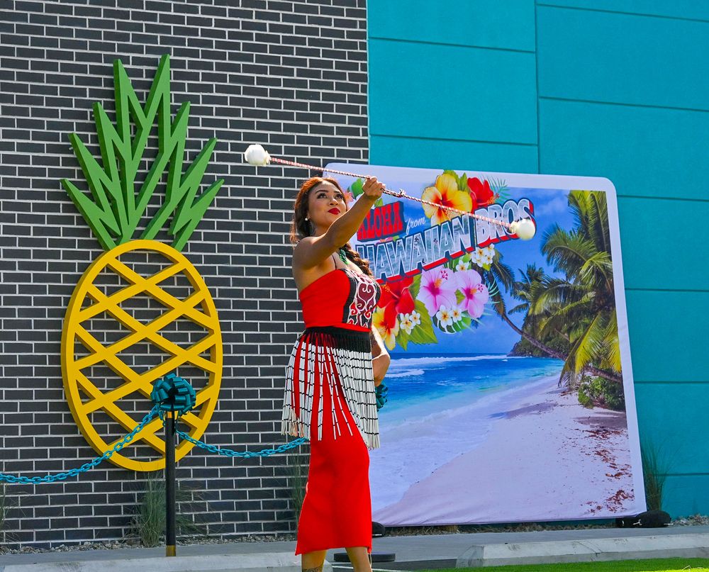 Female performer in red dress juggles white object beside pineapple decor at Hawaiian Bros Island Grill opening.