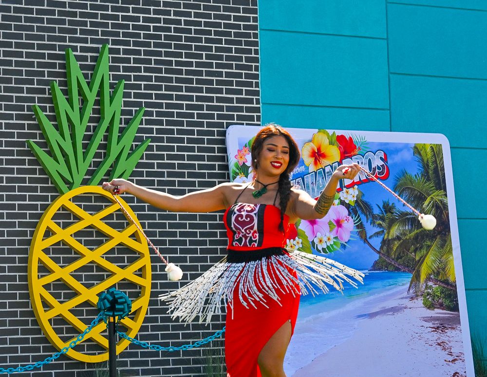 Woman in red Hawaiian dress poses with pineapple decoration at tropical themed outdoor event.