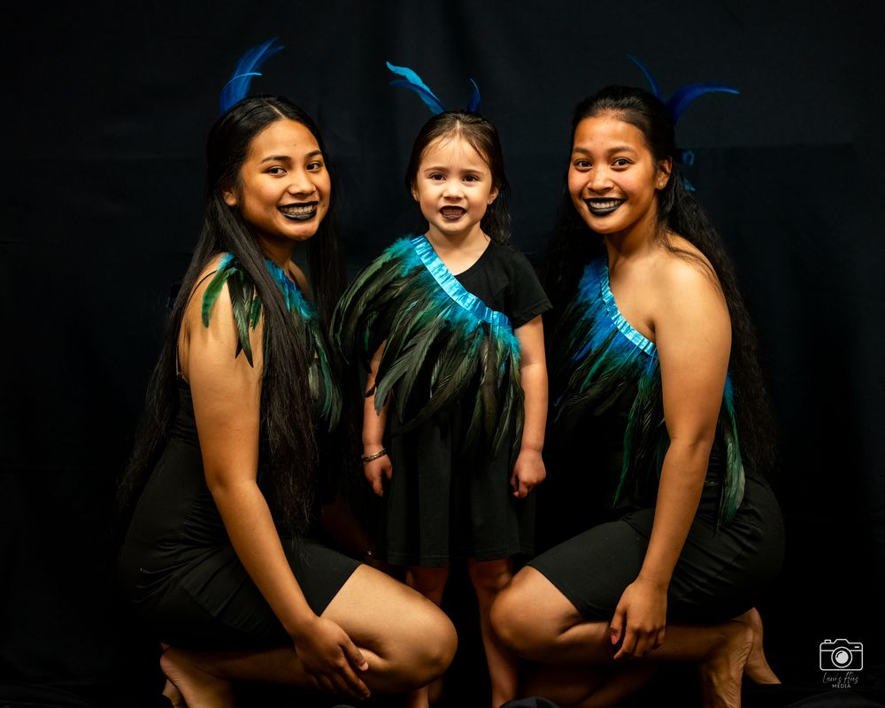 Three graduates in black gowns and blue stoles pose together against a dark background for a celebratory photo shoot.
