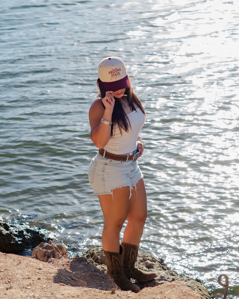 Woman in white top, denim shorts, cowboy boots, and Arizona State cap poses by a shimmering lake.