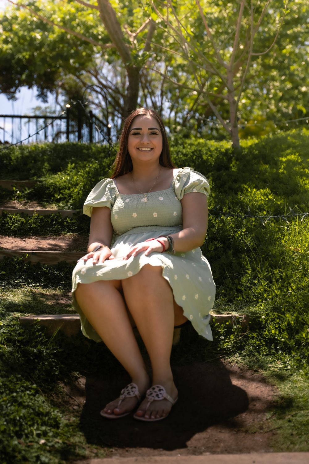 Person in white polka dot dress sits casually on outdoor bench surrounded by greenery.