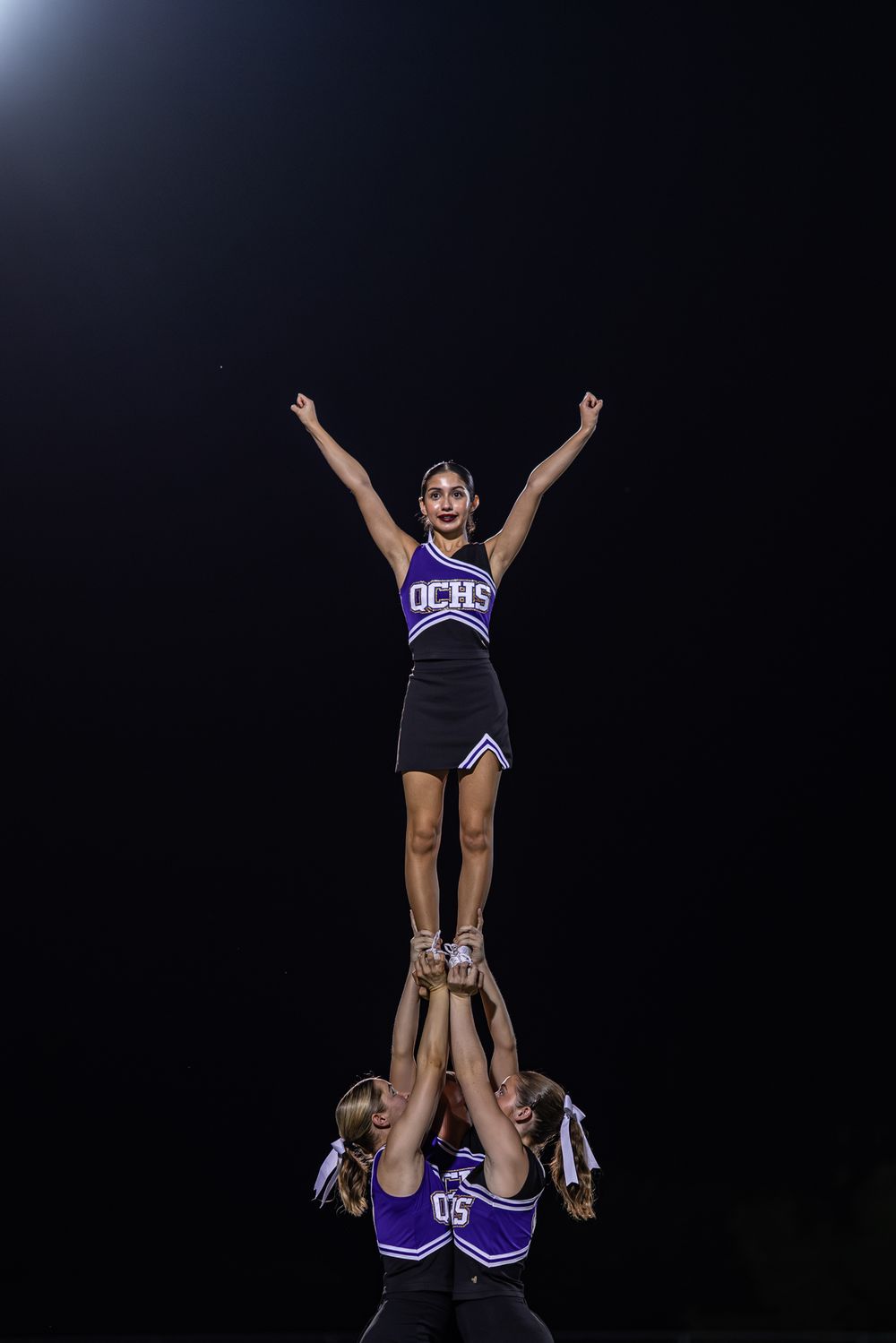 Cheerleaders perform stunts and lifts in purple uniforms against a dark background.