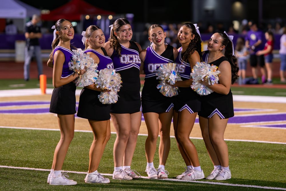 Cheerleaders in purple and black uniforms perform on the sidelines during a sporting event.