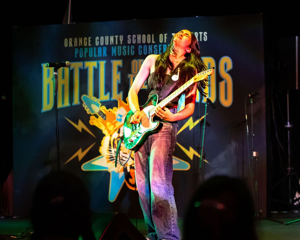 A guitarist performs on stage with colorful lighting and Battle of the Bands signage visible in the background.