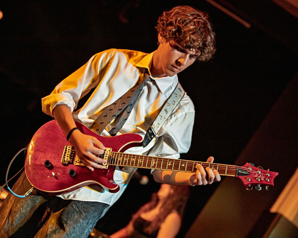 Musician in white shirt plays red electric guitar on stage during live performance under warm lighting.