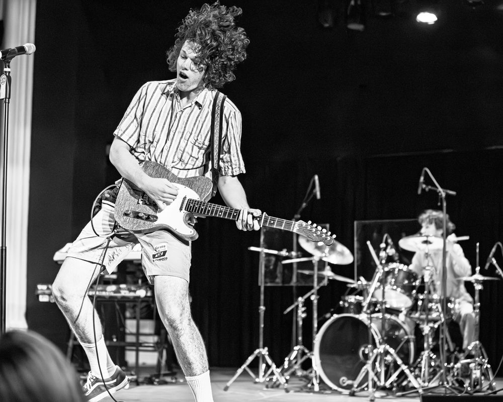 Black and white photo of a rock band performing on stage with electric guitar and drum kit visible in the background.