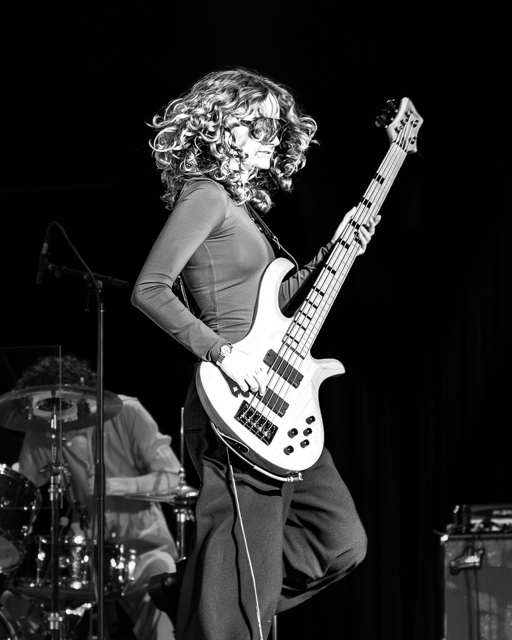 Black and white concert photo showing a bassist performing on stage with a white bass guitar in dramatic lighting.