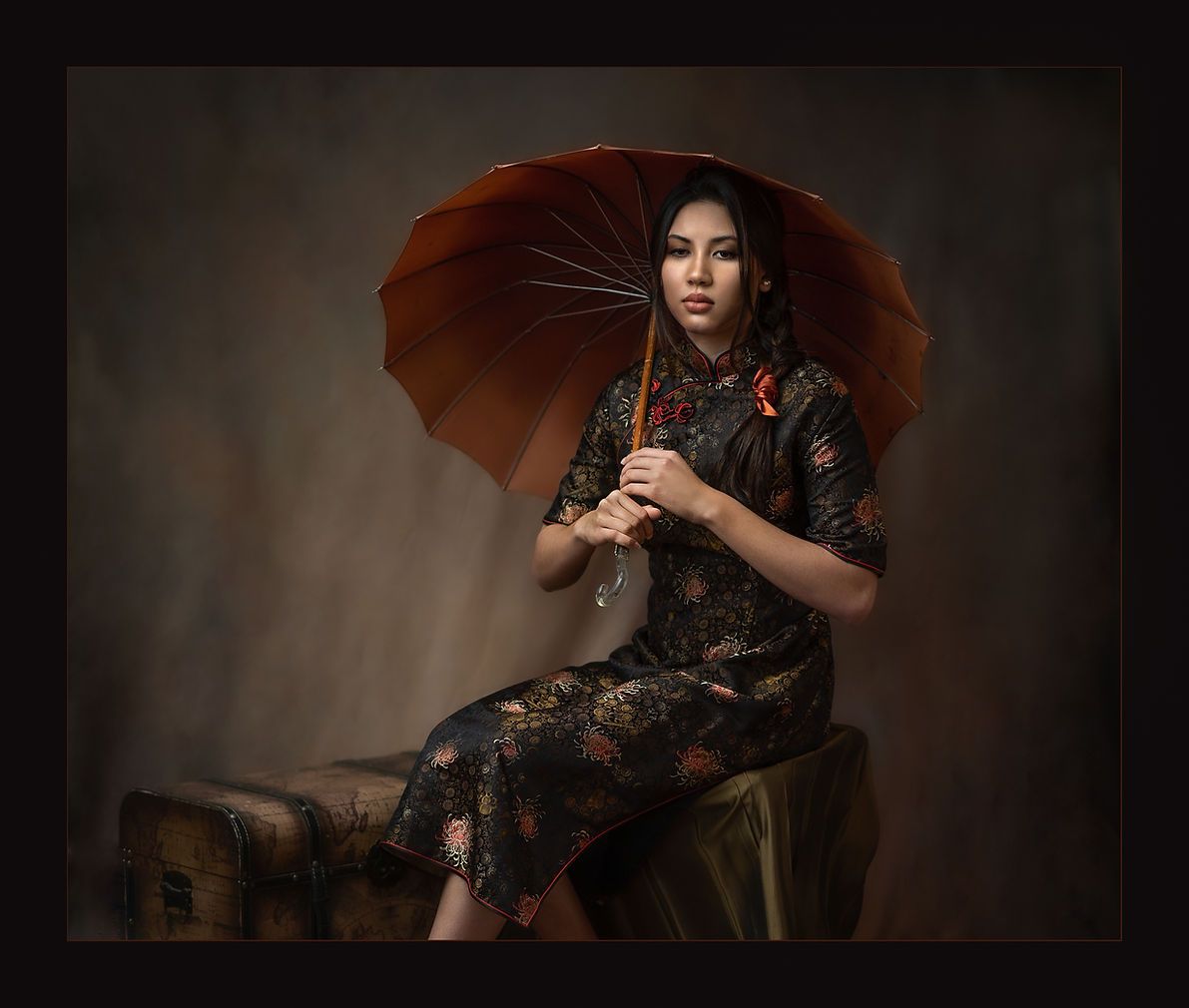 Asian woman in traditional floral qipao holding red umbrella seated on vintage trunk.