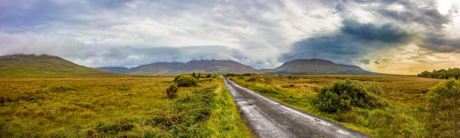 Panoramic view of empty rural road through green Irish countryside with dramatic stormy mountains.